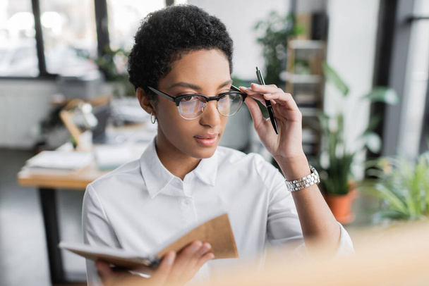 african american businesswoman in white blouse adjusting eyeglasses while holding notepad and pen in office - Foto, Imagem