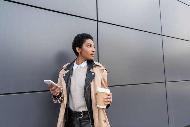 african american businesswoman in stylish trench coat holding paper cup and mobile phone while looking away near grey wall on street - Foto, imagen
