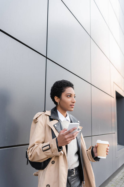 afrikanisch-amerikanische Geschäftsfrau in beigem Trenchcoat steht mit Pappbecher und Smartphone neben grauer Wand an der Stadtstraße - Foto, Bild