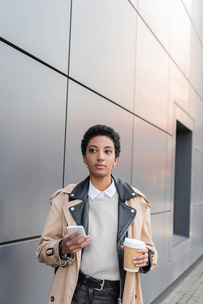 young african american businesswoman in fashionable trench coat holding smartphone and coffee to go near grey wall on urban street - Photo, Image