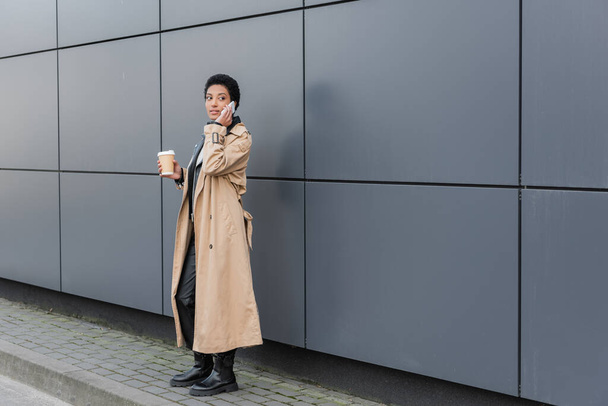 african american businesswoman in beige trench coat standing with takeaway drink and calling on smartphone near grey wall in city - Photo, Image