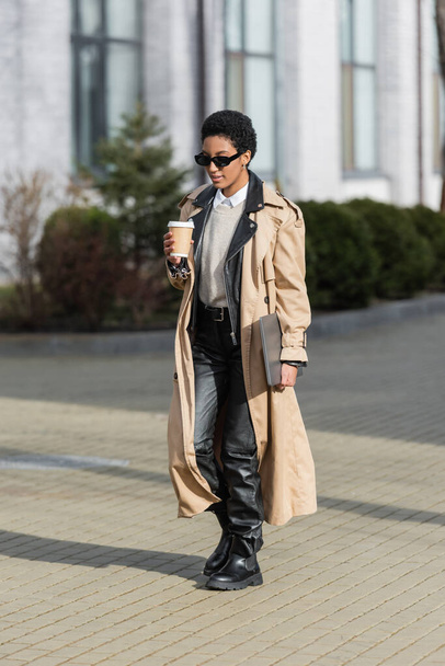 comprimento total de mulher de negócios afro-americana com bebida takeaway e laptop andando em óculos de sol e casaco de trincheira na rua turva - Foto, Imagem