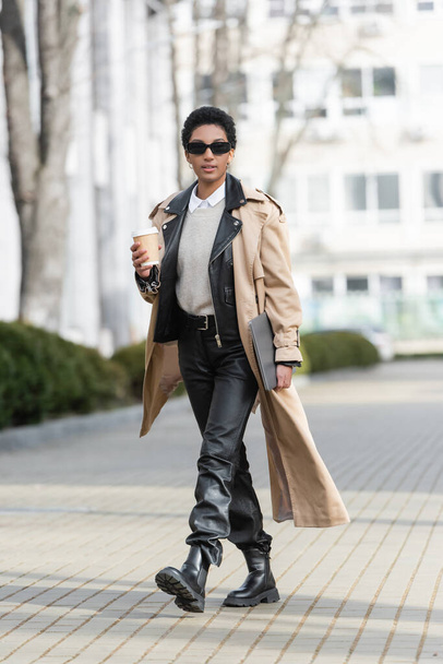full length of fashionable african american businesswoman in sunglasses walking with laptop and takeaway drink on blurred street in city - Photo, image