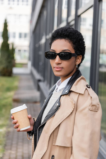 joven mujer de negocios afroamericana en gafas de sol y gabardina sosteniendo café para ir cerca de edificio borroso en la calle - Foto, Imagen