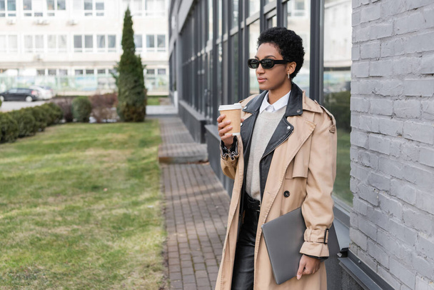 fashionable african american businesswoman in sunglasses standing with laptop and takeaway drink near city building with glass facade - Foto, Imagem