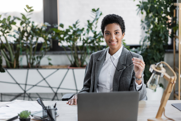 vreugdevolle Afrikaanse Amerikaanse zakenvrouw in grijs pak met bril en kijken naar camera in de buurt van laptop op kantoor - Foto, afbeelding