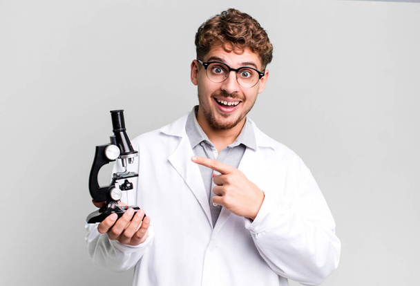 young adult caucasian man looking excited and surprised pointing to the side. scients laboratory student with a microscope concept - Φωτογραφία, εικόνα
