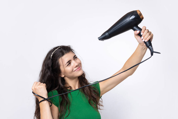 A cute young girl dressed in a green top dries her beautiful long silky hair with a hair dryer against a white background. - Фото, изображение