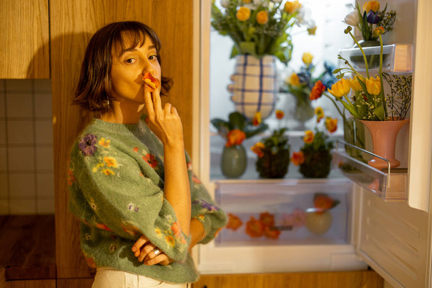 Carina donna apre frigorifero pieno di fiori freschi, in piedi sulla cucina di casa. Concetto di fiori commestibili e dieta di fiore per bellezza - Foto, immagini
