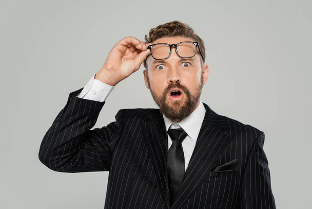 shocked businessman in formal wear adjusting glasses and looking at camera isolated on grey  - Фото, изображение
