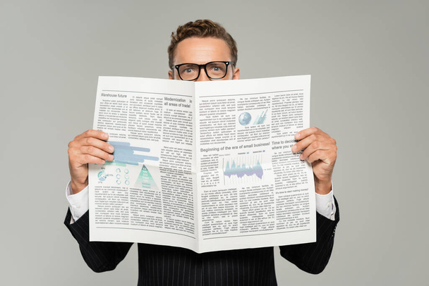 businessman in glasses looking at camera while covering face with newspaper isolated on grey  - 写真・画像