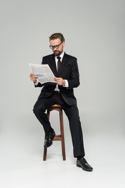 businessman in suit and glasses reading newspaper while sitting on high chair isolated on grey  - Foto, immagini