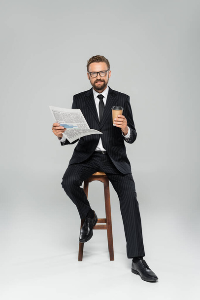 full length of happy businessman in suit and glasses holding newspaper while sitting with paper cup on grey  - Fotografie, Obrázek