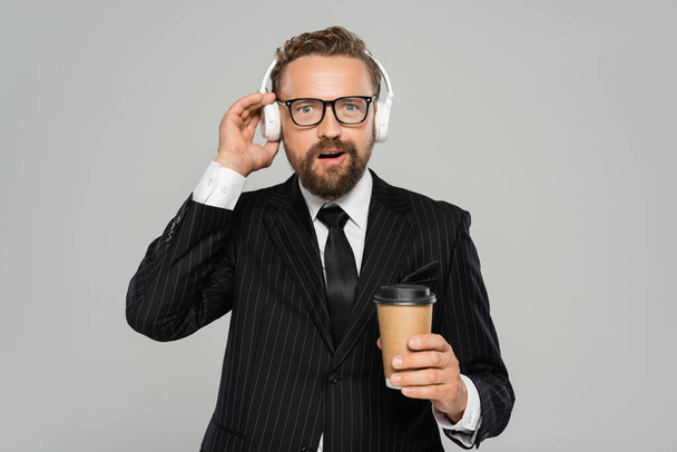 užaslý podnikatel v brýlích a obleku drží papírový pohár při poslechu hudby izolované na šedé  - Fotografie, Obrázek