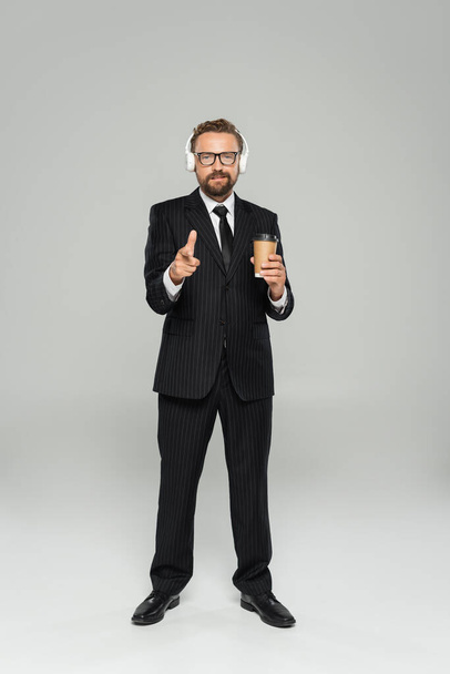 full length of happy businessman in glasses and suit holding paper cup and pointing with finger on grey  - Φωτογραφία, εικόνα