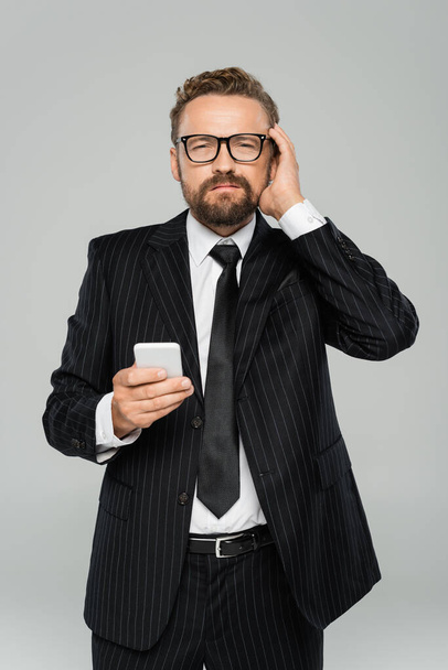 bearded businessman in glasses and suit having headache while holding smartphone isolated on grey    - Foto, imagen