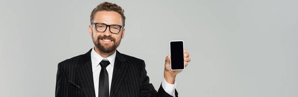 hombre de negocios alegre en traje y gafas que muestran teléfono inteligente con pantalla en blanco aislado en gris, bandera  - Foto, Imagen