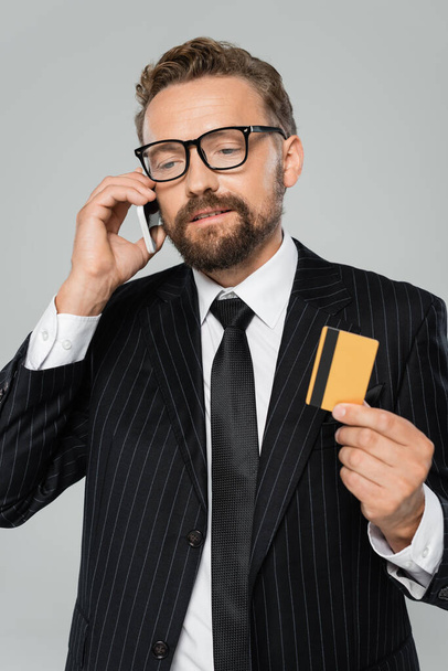 bearded businessman in suit and glasses talking on smartphone and holding credit card isolated on grey  - Foto, Imagen