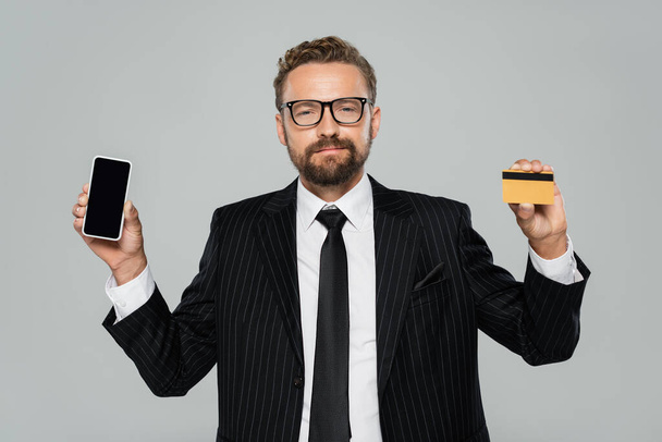 Takım elbiseli sakallı işadamı ve elinde akıllı telefon ve kredi kartı olan gözlüklü adam griden izole edilmiş.  - Fotoğraf, Görsel