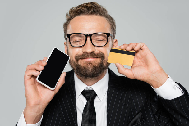happy businessman in suit and glasses holding smartphone and credit card isolated on grey - Fotó, kép