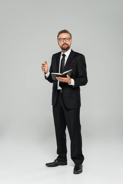 full length of well dressed businessman in glasses and formal wear holding notebook on grey  - Фото, изображение