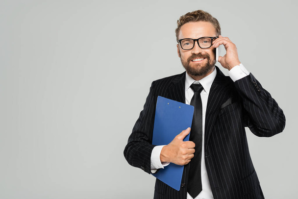 happy businessman in glasses holding clipboard and talking on smartphone isolated on grey  - Фото, зображення