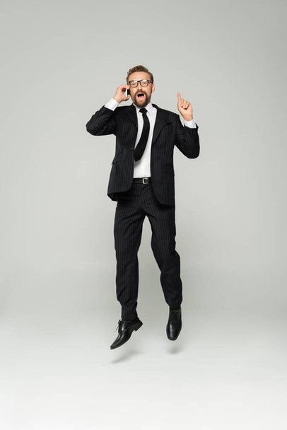 full length of excited businessman in glasses talking on smartphone and pointing up while jumping on grey  - Foto, imagen