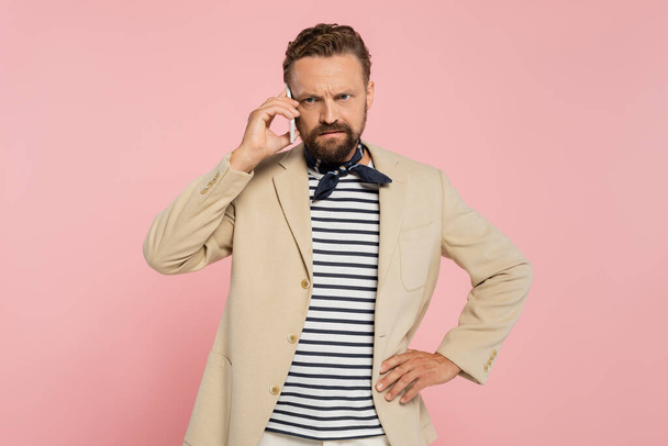Unzufriedener französischer Mann in Blazer und Halstuch spricht auf Smartphone, während er mit der Hand an der Hüfte isoliert auf rosa steht  - Foto, Bild