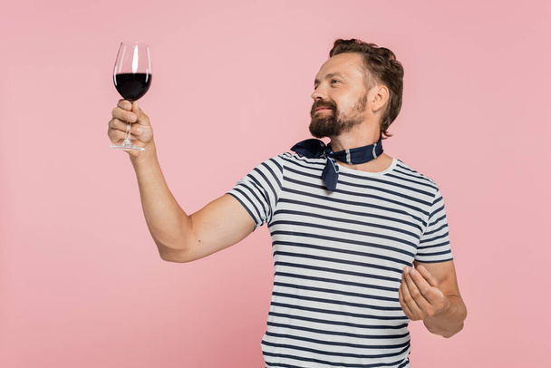 wesoły mężczyzna w pasiastej koszulce trzymając szklankę czerwonego francuskiego wina odizolowanego na różowo  - Zdjęcie, obraz