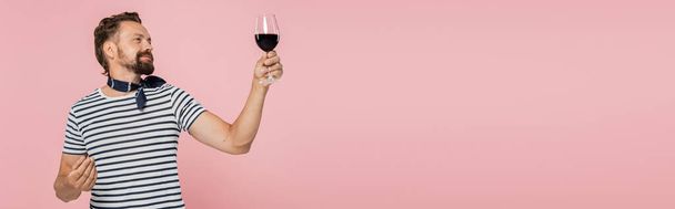 homme heureux en t-shirt rayé tenant un verre de vin rouge français isolé sur rose, bannière  - Photo, image