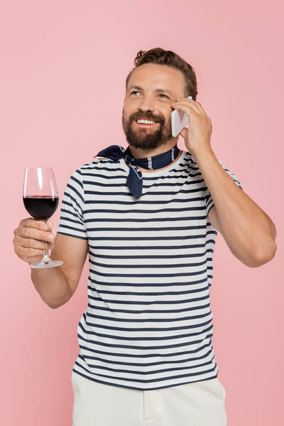 fröhlicher Mann im gestreiften T-Shirt, der ein Glas französischen Rotwein in der Hand hält und isoliert auf einem Smartphone in rosa spricht  - Foto, Bild