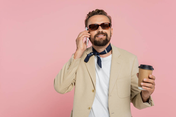щасливий французький чоловік в шарфі і сонцезахисних окулярах тримає каву, щоб піти і розмовляти на смартфоні ізольовано на рожевому
  - Фото, зображення