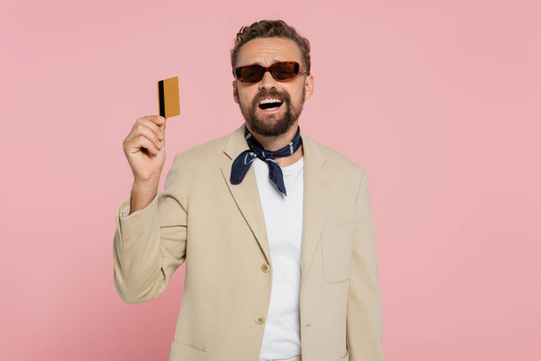 excited man in stylish sunglasses and neck scarf holding credit card isolated on pink  - Photo, Image