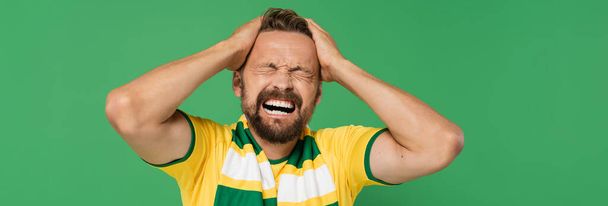 эмоциональный фанат спорта в полосатом шарфе и желтой футболке дергая его волосы изолированы на зеленый, баннер  - Фото, изображение