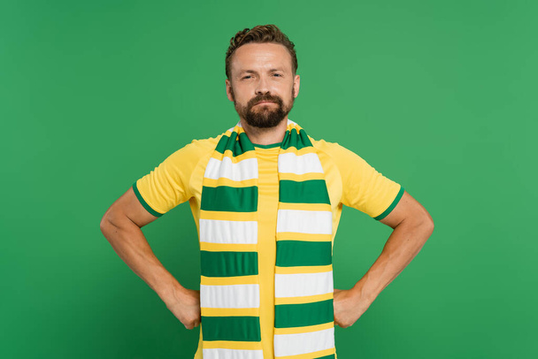 abanico de fútbol barbudo en bufanda a rayas y camiseta amarilla de pie con las manos en las caderas aisladas en verde  - Foto, Imagen