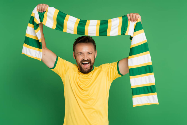 emocionado fanático del fútbol en camiseta amarilla sosteniendo bufanda a rayas y mirando a la cámara aislada en verde  - Foto, imagen