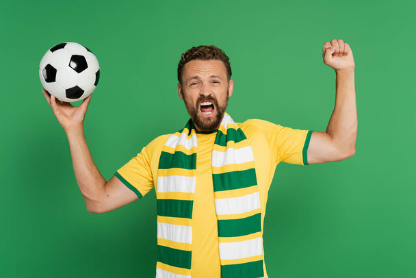 emotional soccer fan in striped scarf holding football and rejoicing isolated on green  - Foto, Imagem