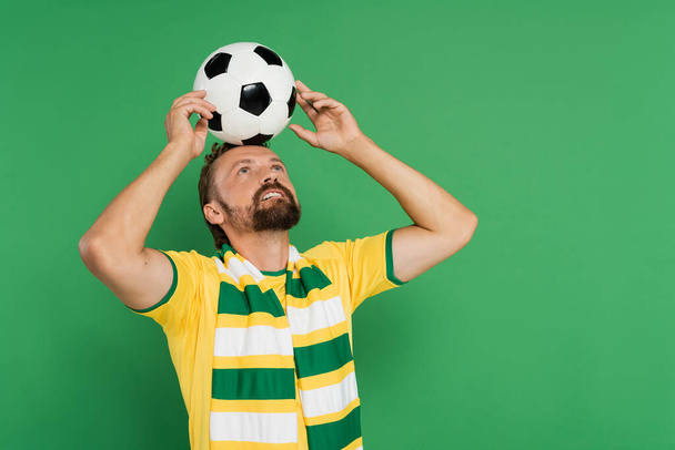 бородатый футбольный фанат в полосатом шарфе и желтой футболке держа футбол на голове изолированы на зеленый  - Фото, изображение