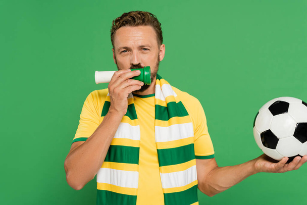 бородатый спортивный болельщик держит футбол и трубит в рог, в то время как приветствия изолированы на зеленый  - Фото, изображение