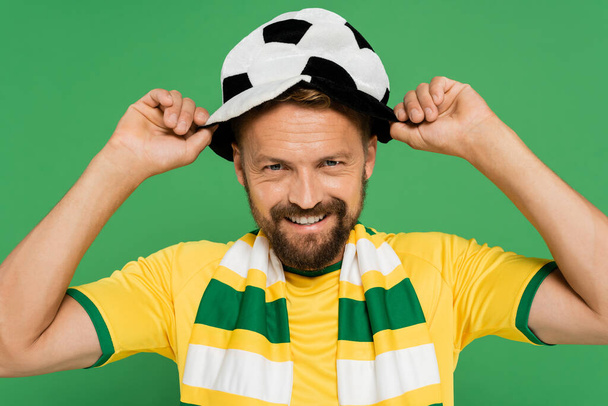 szczęśliwy brodaty mężczyzna w paski szalik noszenie kapelusz kibica piłki nożnej izolowane na zielono  - Zdjęcie, obraz