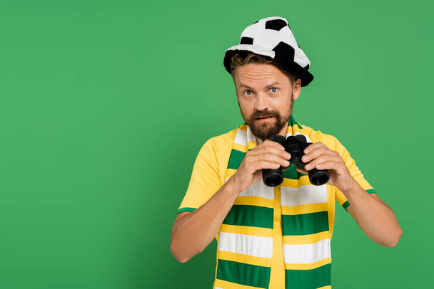 bearded man in fan hat and striped scarf holding binoculars during football match isolated on green  - Photo, Image