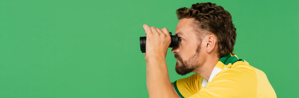 boční pohled na vousatého muže v pruhovaném šátku s dalekohledem při fotbalovém zápase izolovaném na zelené, transparentní  - Fotografie, Obrázek