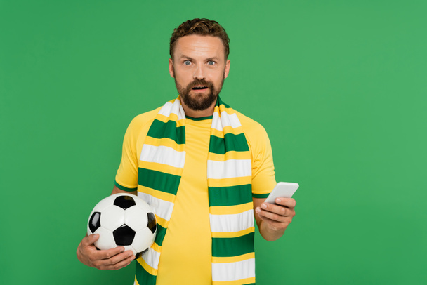 Verbijsterde man in gestreepte sjaal houden voetbal en mobiele telefoon geïsoleerd op groen - Foto, afbeelding