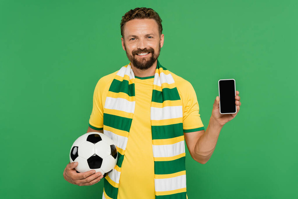 fröhlicher Mann mit gestreiftem Schal, der Fußball und Smartphone mit leerem Bildschirm auf Grün hält - Foto, Bild