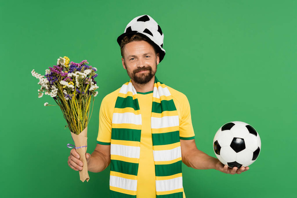 bärtiger Mann mit Sportfan-Mütze, Fußball und Wildblumen vereinzelt auf grünem Grund  - Foto, Bild
