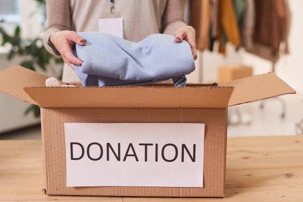 Unrecognizable volunteer standing at table putting clean clothes into cardboard donation box for charity - 写真・画像