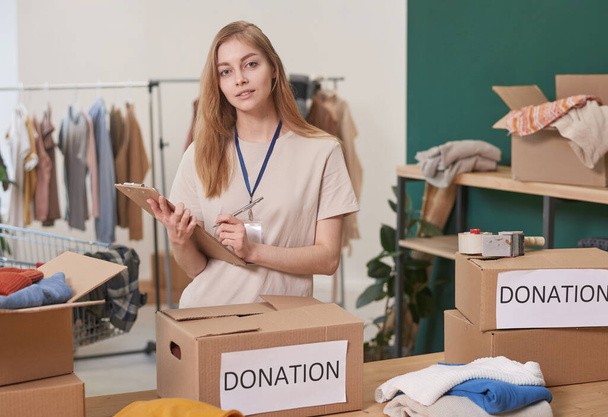 Modern young woman with long hair working in clothes charity organization making list of donation boxes looking at camera - Fotografie, Obrázek