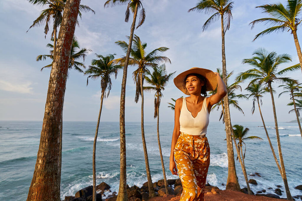 Young african female model posing in colorful clothes at tropical setting at sunrise. Black woman against exotic scenery at dawn. Multiracial dark-skinned model poses in front of palm trees at sunset - Photo, Image