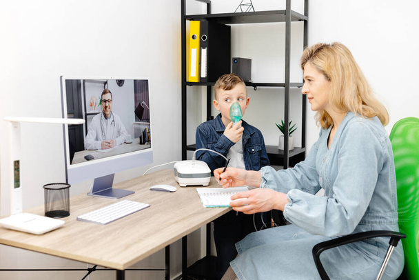 Caucasian little boy making inhalation with nebulizer at home. A young mother helping her son to inhale with a nebulizer at an online meeting with a doctor. - Photo, Image