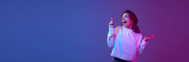 Excited, happy, positive young girl pointing with finger up against gradient blue purple studio background in neon light. Information. Concept of emotions, youth, lifestyle. Banner. Copy space for ad - Foto, immagini